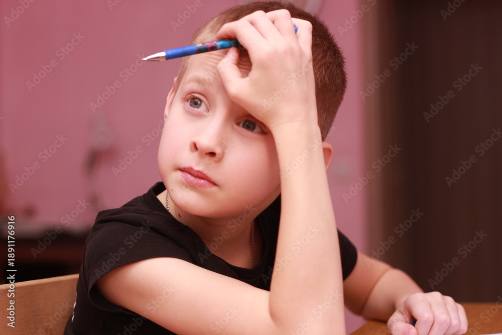 Obraz premium The middle plan of a teenager sitting at a table at home: he leans his elbow on the table and holds a pen to his temple, as if he is considering a task or pauses in his work. It is suitable for illust