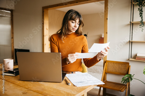 Woman managing finances, checking documents, and using laptop at wooden desk