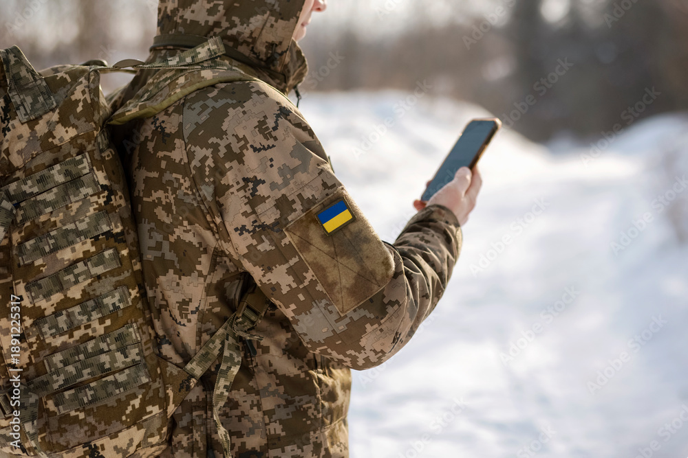 Fototapeta premium Military Soldier with Backpack Using Phone in Winter. Military Navigation Tactical NAV Smart Digital in Ukrainian Soldier Hands.