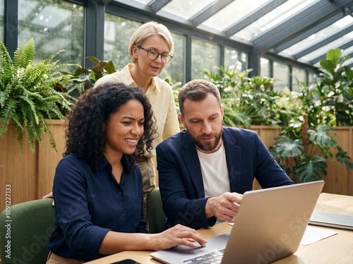 Diverse group of coworkers collaborating on laptop at wooden table in sunny greenhouse workspace