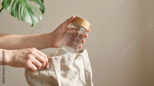 Hands placing a reusable glass water bottle with a wooden lid into a fabric tote bag, promoting eco-friendly lifestyle.