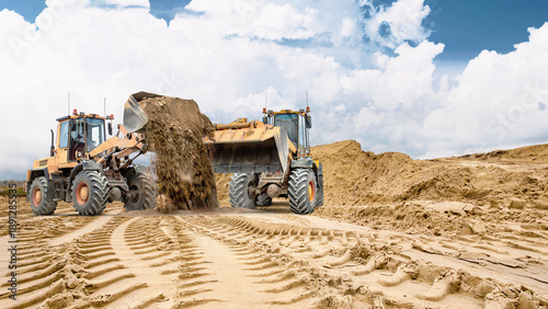 Two wheel loaders. Heavy equipment is working on sandy soil at a construction site. Track marks are visible in the sand, and a large pile of sand is visible in the background