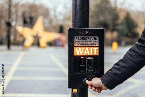 Pedestrian pressing WAIT button at crosswalk on street with holiday lights in the background.