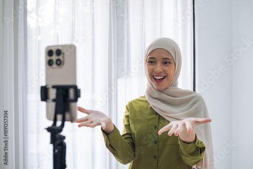 A cheerful hijab-wearing woman records a video with a smartphone on a tripod indoors, showcasing modern lifestyle, content creation, and online connection in a bright, casual home setting.