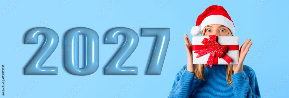 Obraz premium A person wearing a red Santa hat holds a gift box with a ribbon in front of their face. Large numbers display 2027 in blue, creating a festive scene for New Year celebrations.