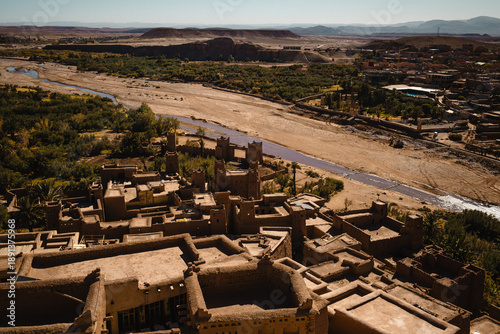 Aerial view of an ancient adobe ksar beside a river oasis, fortified desert settlement with palm groves, mudbrick houses, and arid mountains under clear daylight.