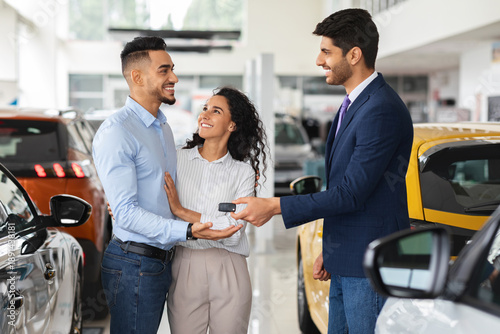Foto Smiling sales manager giving happy middle-eastern young family key from brand new auto, side view