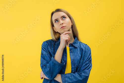 Young pensive woman touching chin, looking up service or advertising text, isolated on yellow studio background.