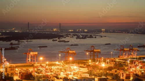 Harbor cranes lift cargo by the river at dusk in Xinzhou, Wuhan, China, with many boats in the water under a colorful sky. Wuhan, China port at dusk with cargo cranes and ships on the Yangtze River