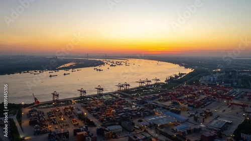 A vast shipping port near the river displays shipping containers and cranes under a soft evening sky over Xinzhou, Wuhan, China. Aerial view of a bustling shipping port in Wuhan, China 