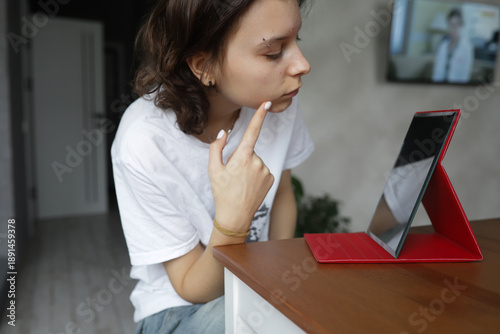 A teenage girl sits at a table in front of a mirror and squeezes pimples while doing facial skin care at home. The scene reflects the typical problems of adolescence, acne, self-esteem, hygiene, and