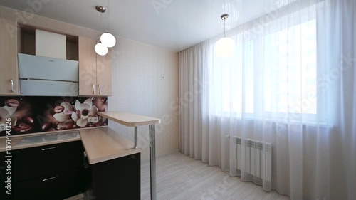 A modern kitchen with black lower cabinets, light upper cabinets, floral backsplash, pendant lights, large window with sheer curtains, and a corner layout — bright and stylish.
