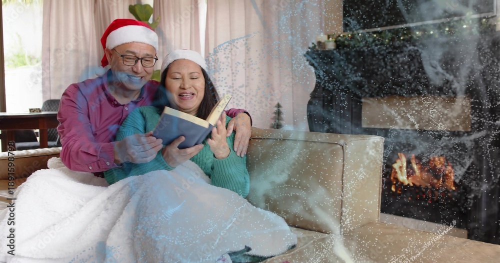 Obraz premium Reading mature couple sitting on living room sofa, wearing Santa hats and sharing open book