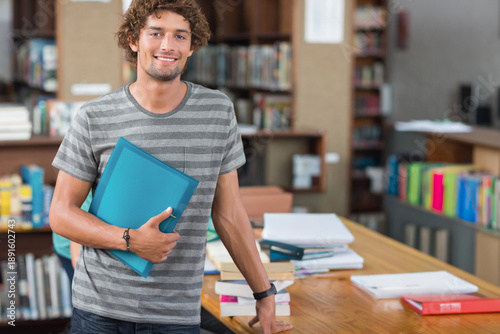 Slika na platnu Adult male standing at wooden study table in library, holding blue binder, near