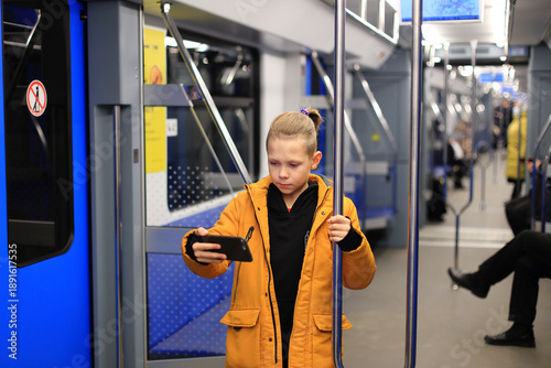 A child in a mustard jacket stands on a metro train, holding a pole and intently using a smartphone, with blue interiors and passengers in the background.