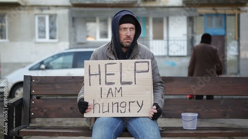 A homeless man in dirty clothes sits on a bench in a city park and asks for alms. Homeless man holds in his hands a paper with the inscription Help, I am hungry.