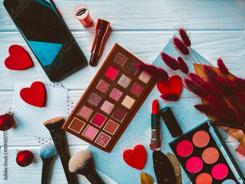 Eyeshadow palette, makeup brushes, red lipstick, hearts, phone, and dried flowers on a white wooden background. Valentine's Day. Red colour.