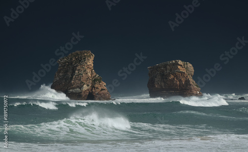Horizontal image of two large rocks battered by the waves of the sea raging in the storm, the waves crashing against the rocks under a dark sky.
