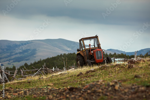 Old, small red and yellow tractor parked on a steep grassy slope in a rural mountain landscape.