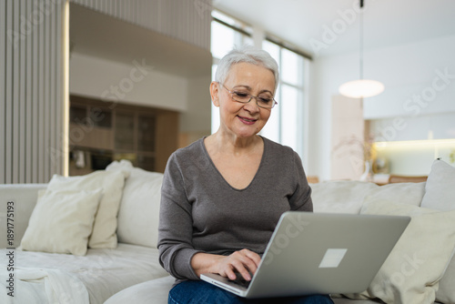 Confident stylish happy middle aged senior woman using laptop at home. Stylish older mature 60s grandmother sitting at couch looking at computer screen typing chatting reading writing email