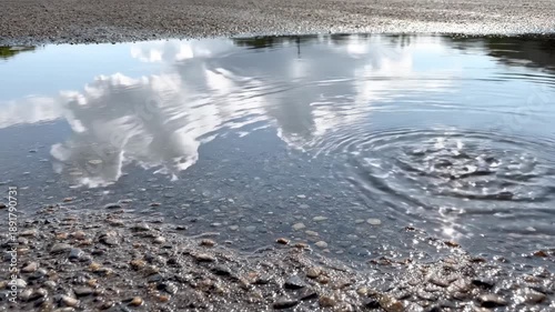 Cloud reflections in puddle. Clouds drifting overhead reflected clearly in a shallow puddle on flat ground, the reflection gently disturbed by faint ripples.