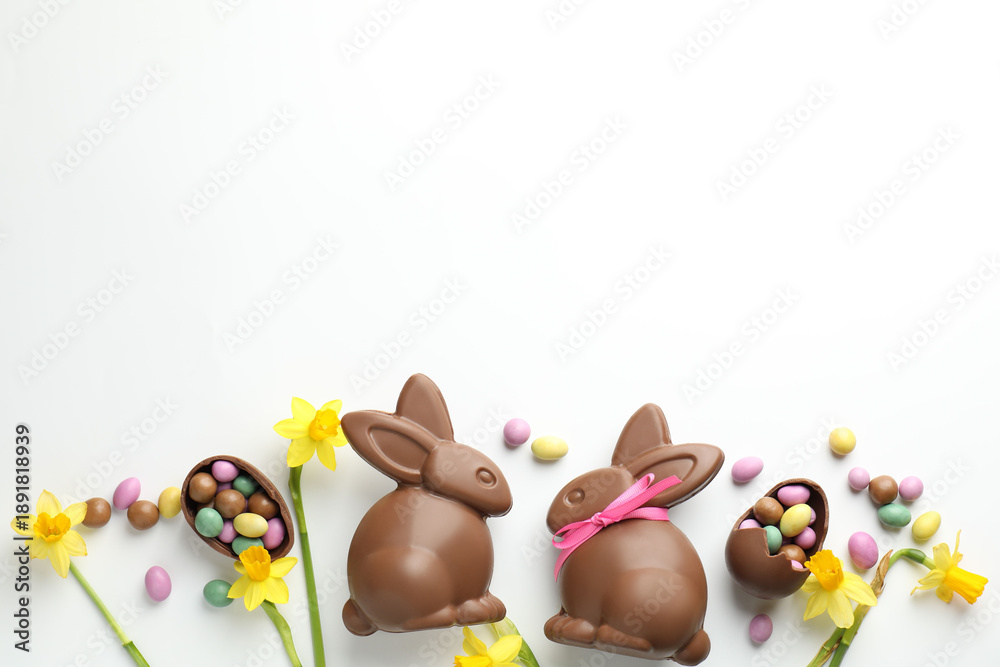 Obraz premium Chocolate Easter bunnies, flowers, ribbon and eggs on white background, flat lay. Space for text