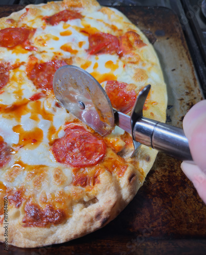Closeup of a hand using a stainless steel, cutting wheel tool to slice into a hot, oven baked, pepperoni pizza.