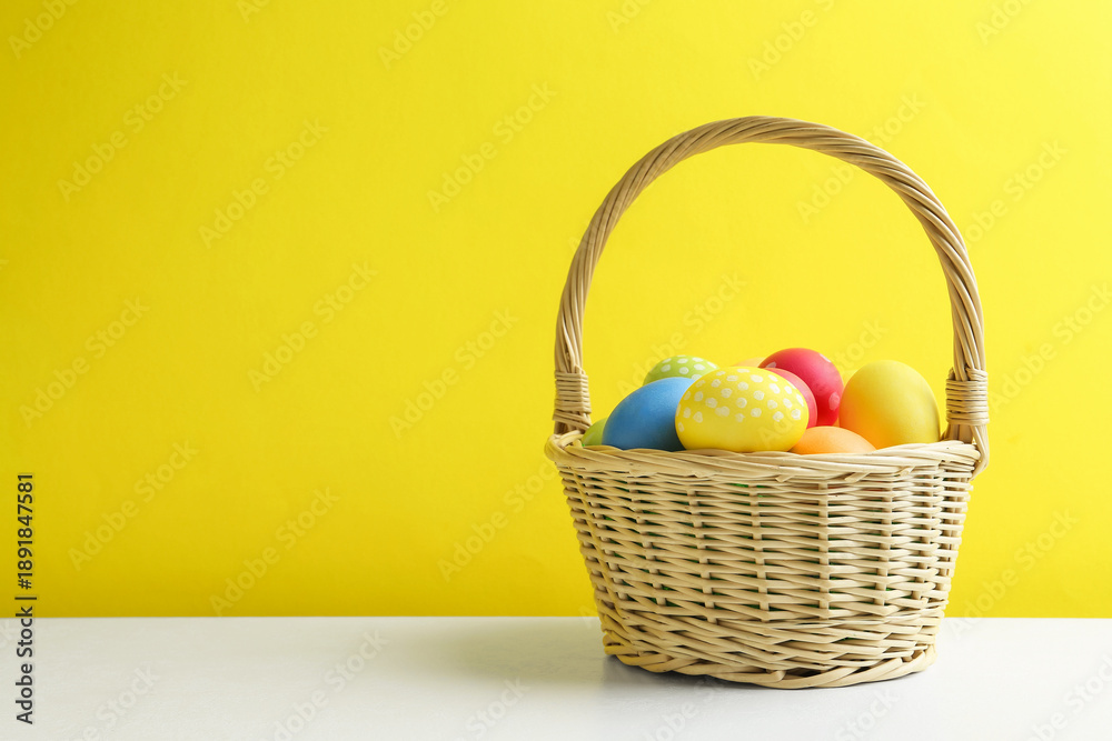 Fototapeta premium Painted Easter eggs in wicker basket on white table against yellow background. Space for text