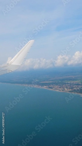 airplane wing remains visible above calm blue coastline sandy beaches compact seaside towns meet inland farmland beneath soft band white clouds aerial view horizon vista air travel 