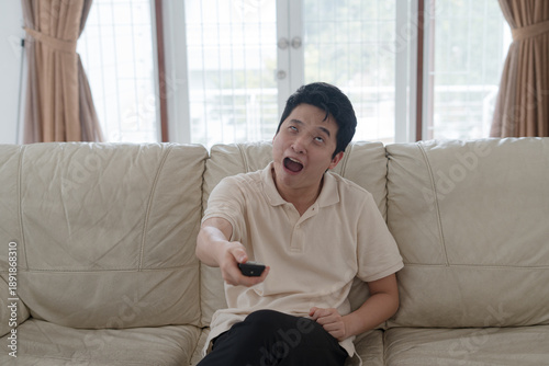Photos Tired young Asian man yawning while holding a remote control on a comfortable so