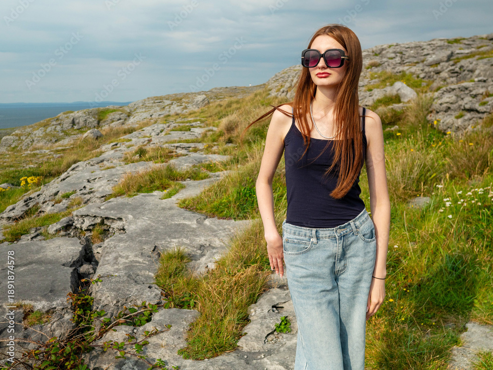 Naklejka premium Young woman enjoys rough rock nature of Burren area of Ireland. Travel and tourism. Warm sunny day. Ocean and Aran island in the background. Popular region with stunning views. Stylish model.