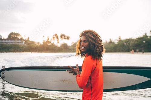 Photography Portrait of black long-haired teen boy with surfboard ready for surfing with sunset backlight