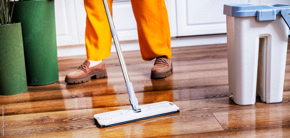 Naklejka premium Professional cleaner washes wooden laminate floor. Woman in orange pants cleans house interior. Household