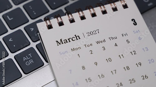 Close-up of a desk calendar showing March 2027 positioned on a laptop keyboard. Camera slowly moves to the right, presenting clear monthly date structure and calm time planning concept in a modern of