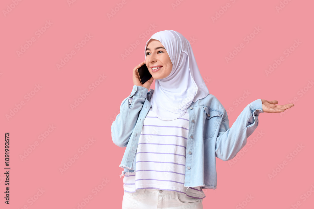 Fototapeta premium Beautiful young Muslim woman in hijab talking by mobile phone on pink background