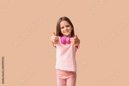 Happy little girl with headphones showing thumbs-up on beige background