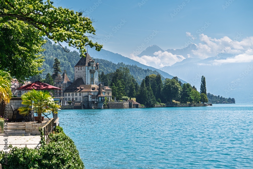 Fototapeta premium Scenic view of Lake Thun featuring Spiez Castle, framed by vibrant greenery and the stunning Swiss Alps. Perfect for travel brochures and tourism promotions, showcasing serene landscapes.