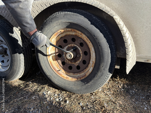 A person in a work glove uses a lug wrench to remove a tire from a rusted rim on a utility trailer. Close-up shot of outdoor maintenance and repair on a gravel surface