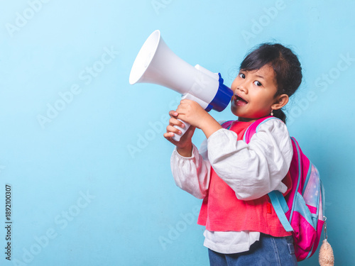 Happy little girl wearing backpack shouting into a megaphone isolated on blue background. Active school child using loud speaker for announcement