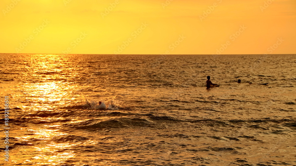 Fototapeta premium A serene summer landscape at sunset featuring the orange silhouette of a man walking along the calm ocean beach as waves reflect the golden horizon and clouds over the sea