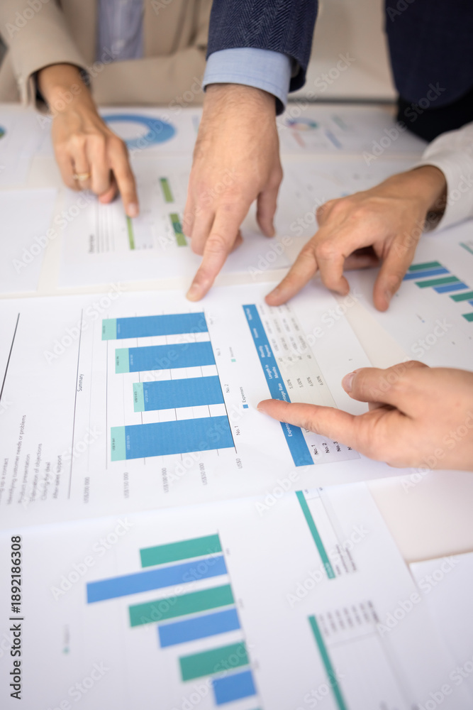 Fototapeta premium Close-up of hands pointing at diverse bars and figures on printed financial charts laid out on conference table, collaborate, analyze data trends, discussing key performance indicators during meeting