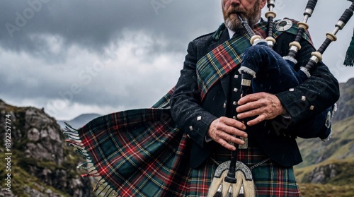 A bearded man in traditional Scottish Highland dress, including a tartan kilt and sash, plays the bagpipes amidst a rugged, misty mountain landscape under a dramatic, cloudy sky.