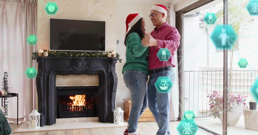 Fototapeta premium Dancing mature couple holding hands in living room, wearing Santa hats, mantel and teal AR icons