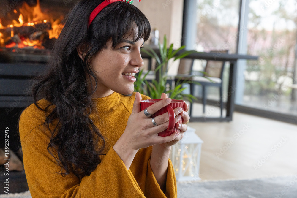 Fototapeta premium Woman wearing mustard sweater and red headband holding mug near fireplace in living room