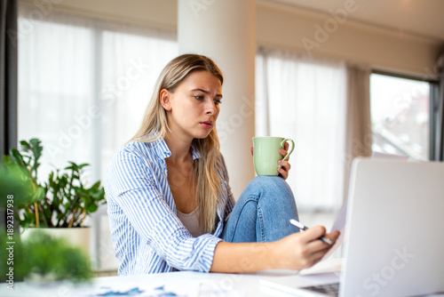 Bilde på lerret Concerned Young Woman Holding Coffee Mug and Reviewing Documents at Home Desk