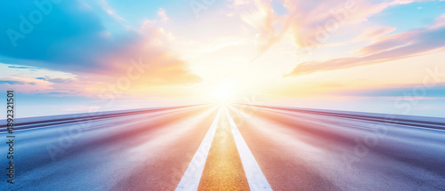 Wide symmetrical road with white markings leading into bright glowing sun against soft blue and pink pastel sky with light clouds
