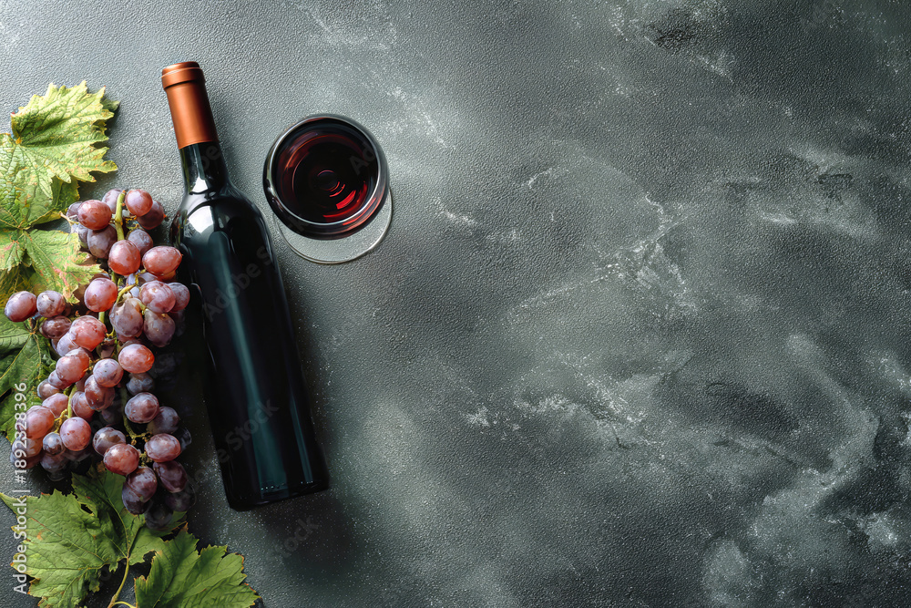 Naklejka premium A wine bottle stands next to a glass filled with red wine and fresh grapes on a dark background. This setup provides a display for vineyard themes or wine promotion