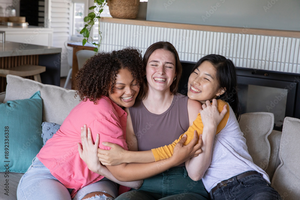 Fototapeta premium Diverse female friends hugging on sofa in home living room, laughing with fireplace backdrop