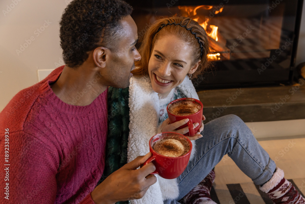 Fototapeta premium Diverse couple sitting on rug before hearth fireplace with red mugs and draped fleece blanket