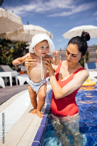 Wallpaper Mural Young woman holding a toddler in a swimming pool, both enjoying a sunny day, with lounge chairs and umbrellas in the background, capturing a joyful family moment Torontodigital.ca